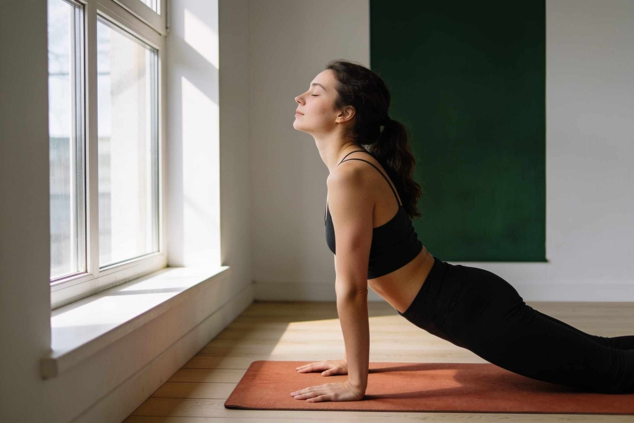 A woman stretches her spine on a mat by a sunny window.
